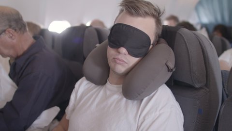 Young man sitting on an airplane sleeping, wearing a neck pillow. Also wearing blindfold