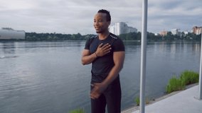Muscular young afro american man in black sporty clothing flexing muscles before workout near the lake in the center of small city. Sportsman is going to do everyday training on nature