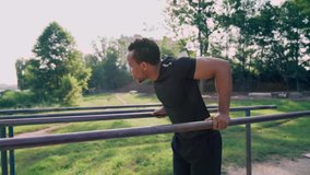 Young male afro american athlete with dark hair in black sports wear doing abdominal exercise on parallel bars in sport ground. Muscular man training outdoors