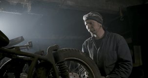 Man blowing dust off the tyre of a vintage motorbike in a workshop or storage facility in dim light