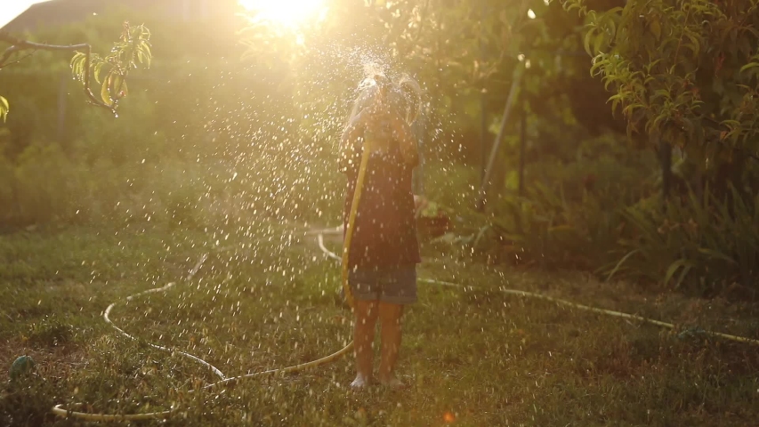 pretty small girl pours water on head from hose against sisters on lush green lawn in summer house backyard at sunlight