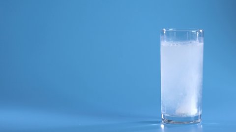Effervescent Tablet Falling In Water. Pill Falls And Dissolves With Bubbles. Effervescent Tablet In Glass Of Water On Blue Background. Shot Of Effervescent Pill Dissolving In A Glass Of Water.