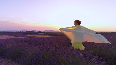 AERIAL, LENS FLARE: Flying around cheerful woman enjoying the sights and scents of idyllic Provence at picturesque summer sunset. Girl walks across a fragrant field of lavender on a sunny evening.