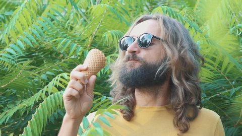 Portrait of long hair bearded musculine man. He licking ice cream like wild cat. Yellow t-shirt and black sunglasses. Green leaves. Be free and wild. Eating tasty chocolate ice cream in waffle cone.