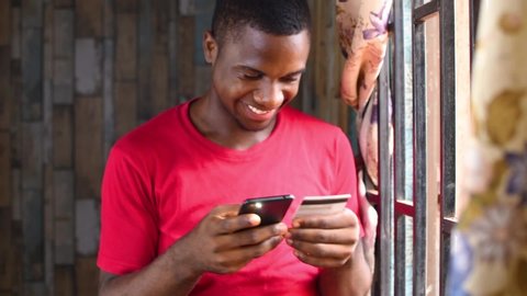 African man holding Bank Credit card Paying Bill's online with his smartphone beside his window.Black man shopping online  for his girlfriend with his mobile phone