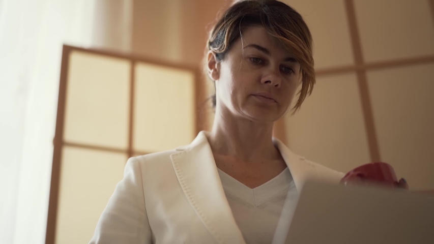 Cute adult caucasian woman drinking coffee and looking at a laptop screen at home. Woman kneads stiff neck. Slow motion. | Shutterstock HD Video #1036096646