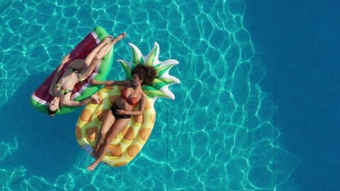Drone view of happy diverse girls sunbathing on colorful air lilo balls in outdoor swimming pool. Aerial shot of multiracial female friends relaxing on floaties in clear blue water of hotel pool