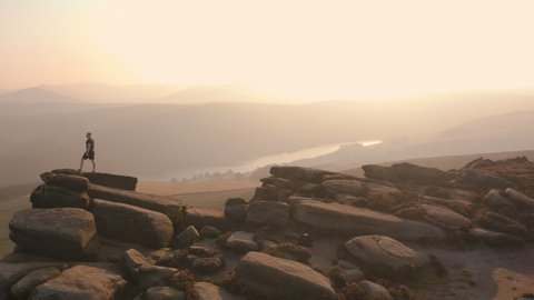 Aerial 4K Drone footage flying around a young white bald male climbing large rocks and boulders whilst hiking along a stunning hillside at sunset. Shot in the Peak District National Park, UK