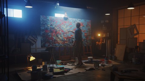 Talented Female Artist Works on Abstract Oil Painting, Using Paint Brush She Creates Modern Masterpiece. Dark and Messy Creative Studio where Large Canvas Stands on Easel Illuminated. Zoom out