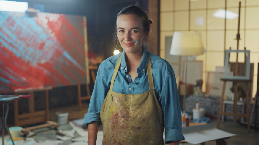 Young Female Artist Dirty with Paint, Wearing Apron, Crosses Arms while Holding Brushes, Looks at the Camera with a Smile. Authentic Creative Studio with Large Canvas. Head and Shoulders Portrait | Shutterstock HD Video #1036107653