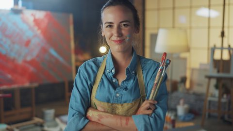 Young Female Artist Dirty with Paint, Wearing Apron, Crosses Arms while Holding Brushes, Looks at the Camera with a Smile. Authentic Creative Studio with Large Canvas. Head and Shoulders Portrait