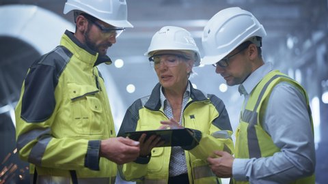 Three Heavy Industry Engineers Stand in Pipe Manufacturing Factory, Use Digital Tablet Computer, Have Discussion. Large Pipe Assembled. Design and Construction of Oil, Gas and Fuels Transport Pipeline