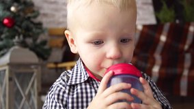 Closeup view of cute little baby boy drinking tea from special children plastic bottle holding it in small hands. Child in Christmas background. Real time 4k video footage.