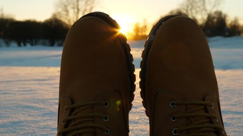 Toes of winter boots against setting sun, man stretch out legs on snow, close shot of warm leather winter shoes. Cold season at European country, blurred city park seen on background