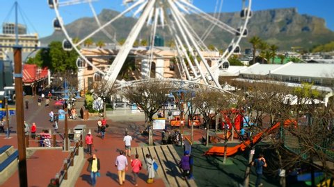 CAPE TOWN, SOUTH AFRICA, CIRCA AUGUST 2019, Elevated tilt shift view Cape town V&A Waterfront Ferris wheel rotating with people walking with Table Mountain blue skyline background
