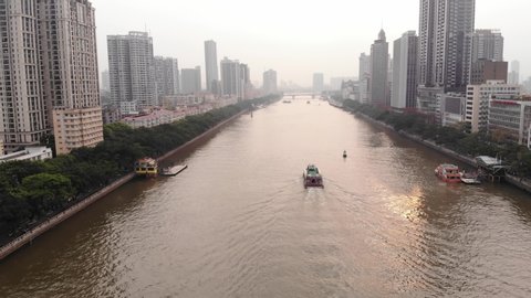 One vessel sail at Pearl River, aerial wide angle shot, tall hotels and residential buildings on shores, Renmin Bridge seen at distance ahead. Green trees grown along water, light mist in air