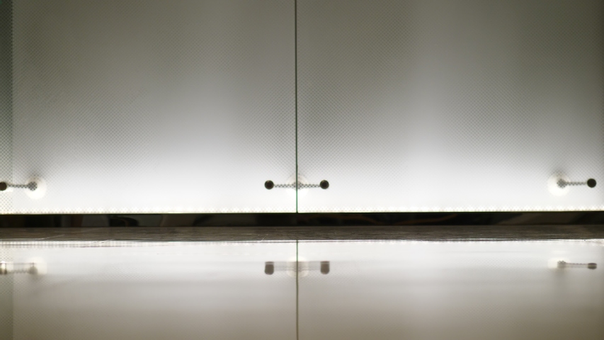 Woman walk at passage with wheeled suitcase, low half slow motion shot. White glass wall illuminated from bottom on background. Slender legs of young adult lady, she wear short skirt and white shirt
