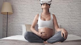 Video of beautiful young pregnant woman using virtual reality glasses while sitting on the bed at home.