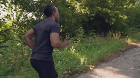 Muscular african guy in black sports wear enjoying fesh air while running along the road with green trees. Fitness, healthy, lifestyle concept