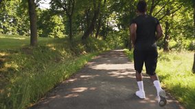 Young muscular afro american athlete wearing black t-shirt, shorts and white sneakers running on asphalt path among green trees during summer sunny days.