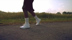 Close up of afro american muscular man's legs in white sneakers and black shorts that is running on asphalt path among green field. Young guy training regularly to be in good body shape