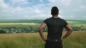 Active muscular african man with dark hair wearing black t-shirt, keeping his hands on hips and looking at picturesque view from the top of mountain. Concept of sport and beauty of nature