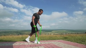 Fitness sports african man with dark hair wearing black t-shirt, shorts and white sneakers exercising with green elastic rubber band on the ground plot of the hill covered by pavements