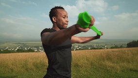 Side view of young muscular afro american man in black t-shirt working out with green rubber expander with background of green field. Handsome guy building muscles in the upper and lower arms