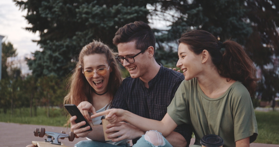 Happy smiling teenage friends laughing outside at something in smartphone or mobile phone. Three young multiracial people spending time together. Friendship, communication, youth and lifestyle concept Royalty-Free Stock Footage #1036137953