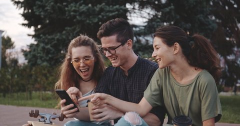 Happy smiling teenage friends laughing outside at something in smartphone or mobile phone. Three young multiracial people spending time together. Friendship, communication, youth and lifestyle concept