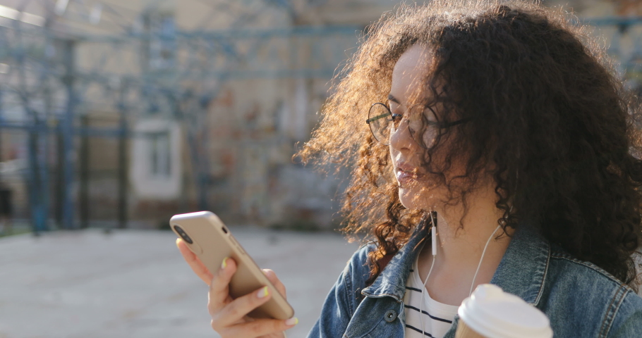Close up of the young Caucasian attractive girl in glasses and with curly hair tapping and texting on the smartphone in the slums at the sport court.