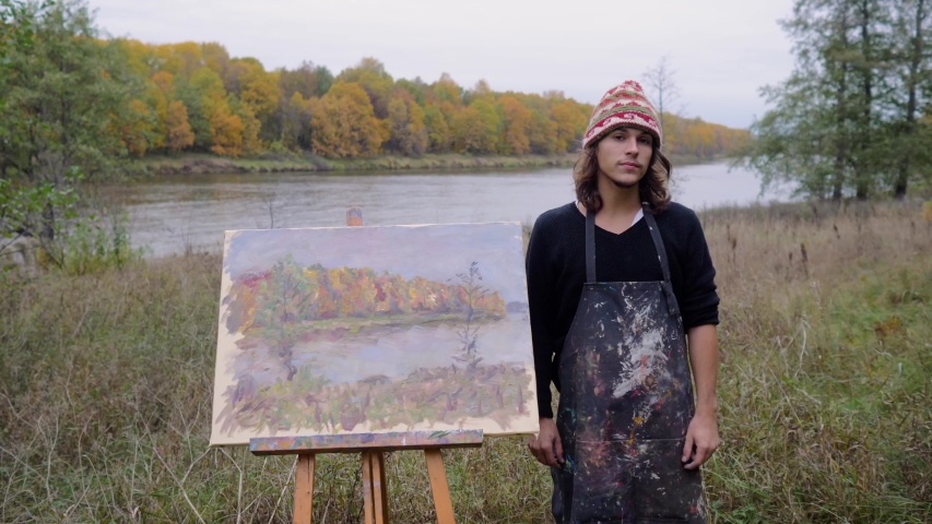 A young man stands next to his painting on the river Bank. The artist next to his painting. Young man draws landscape on the river Bank in autumn 4K