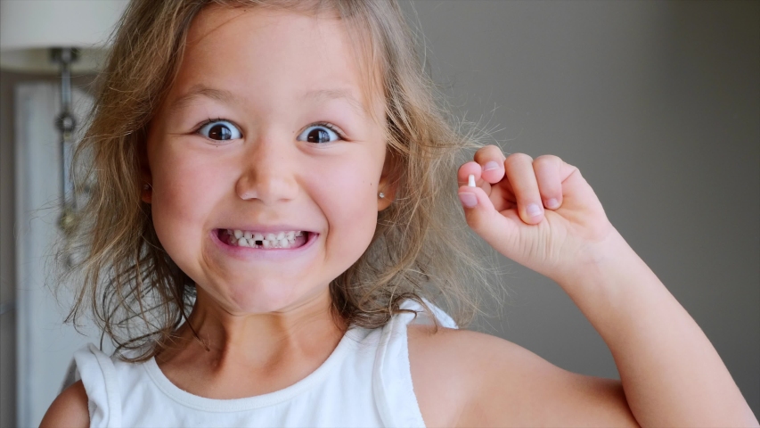 Portrait of cute little child girl is rejoices and showing her lost milk tooth and smiling to camera of toothless mouth while she standing in bedroom. Happy baby holding her fallen tooth in hand | Shutterstock HD Video #1036143071