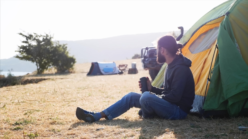 Young bearded hipster man sits on the ground next to touristic tent at expedition campsite on lake shore and drinking tea or coffee from thermos bottle at morning. Side view with backlit