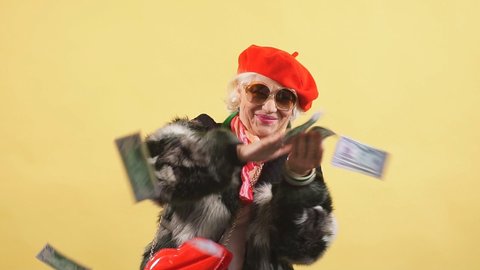Rich elegant woman in fur coat and red cap throwing money, spending money on useless thing, isolated yellow background. studio shot