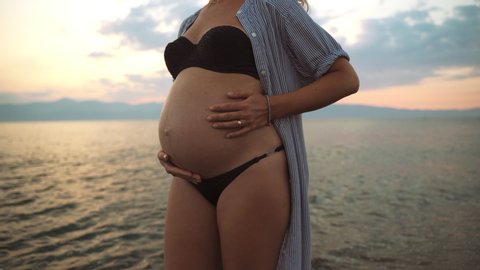Beautiful woman's pregnancy at the sea sunset background. Happy woman in expectation caressing and embracing lovely her tiny belly with her hands. Healthy pregnancy of happy mother. Pregnant tummy