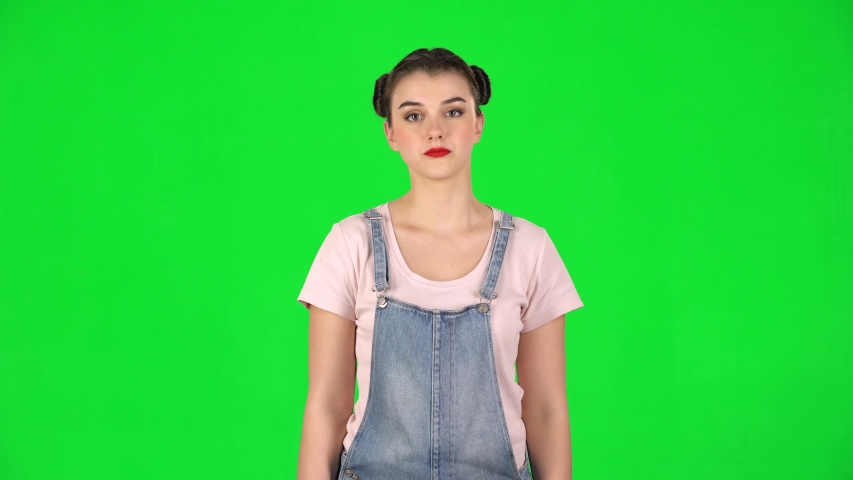 Girl takes out a small box from the packages and is very happy on a green screen in the studio. Girl with two hair-buns and red lips, wearing denim overalls and pink t-shirt.