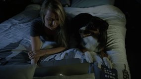 Young woman in bed with her dog streaming movies on her laptop