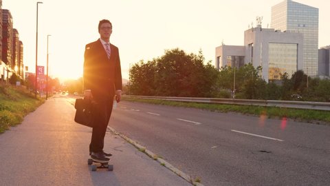 SLOW MOTION, LENS FLARE: Cool sporty businessman going home from work on his e-skateboard. Cheerful yuppie cruising through the city on a sunny summer evening. Happy young man enjoying his ride home.