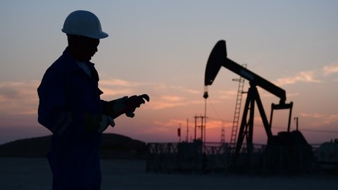 Silhouette of a technical oilfield worker monitoring crude oil pump at sunset.