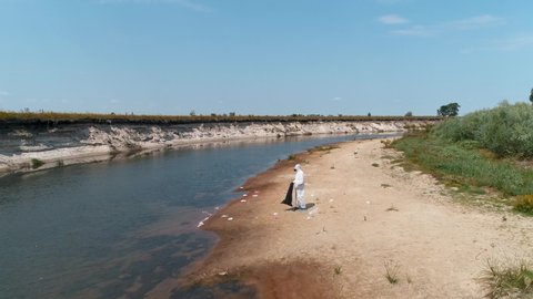 Aerial view of a man in a protective suit and a respirator who collects plastic trash on the banks of a dry and polluted river with a bird on background. Ecological catastrophy. 4K drone footage.