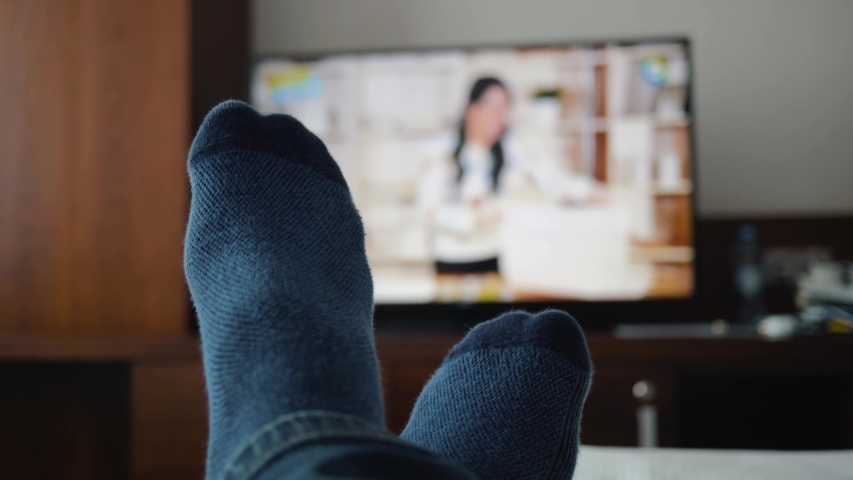 Legs in blue socks lie in front of the TV. Close-up on his feet. Cozy atmosphere of the room. A man watches ads on smart tv. Watching TV. Rest on a day off. Point of view TV watching. Man watch movie Royalty-Free Stock Footage #1036173431