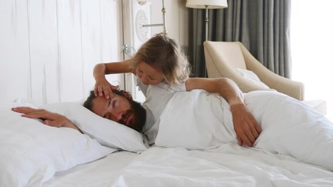 Little cute daughter comes to sleeping father at morning, and opens his eyes by fingers for wakes him. Dad covering his head by pillow. Family at home concept.