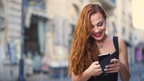 Young fashionable redhead woman with smartphone text messaging and laughing in city in summer