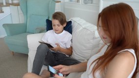 Son emotionally playing on tablet at video game . Mother using her smartphone
