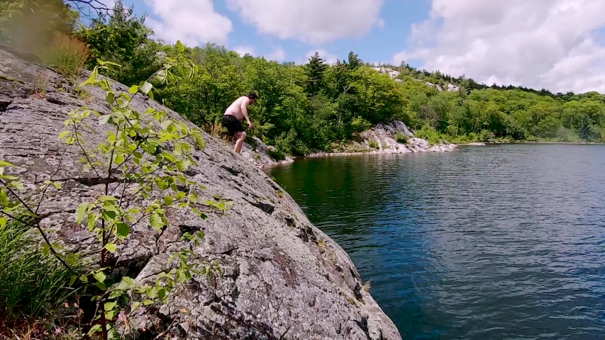 Young White Man Jumps Off Rock Into Blue Lake in Slow Motion, Wide Pan. Male Swimmer Waves Arms and makes Splash as he Submerges in Cool Refreshing Water in Wild Outdoors in Summer Sunlight in Slo Mo
