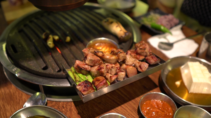3cooking Samgyeopsal on a Korean BBQ Grill. Close up of cooked Pork Belly and Pork Chop next to grill. A piece of the delicious meat is dipped in the sizzling hot dipping sauce.