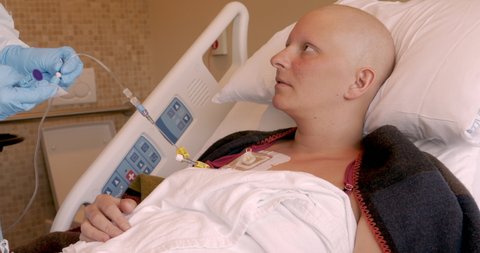 Nurse connecting an infusion connector to an IV drip as part of a central venous port for chemotherapy using sterile techniques for a young female cancer patient lying in a hospital bed