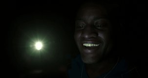 BCU dark skinned man talking on video call through cellphone at night, looks from screen to viewer and smiles. Hand-held 4K with lens flares.