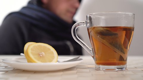 Caucasian man blowing nose because of flu or cold. Drinking black tea with lemon as a natural remedy. Autumn or fall season disease or illness.